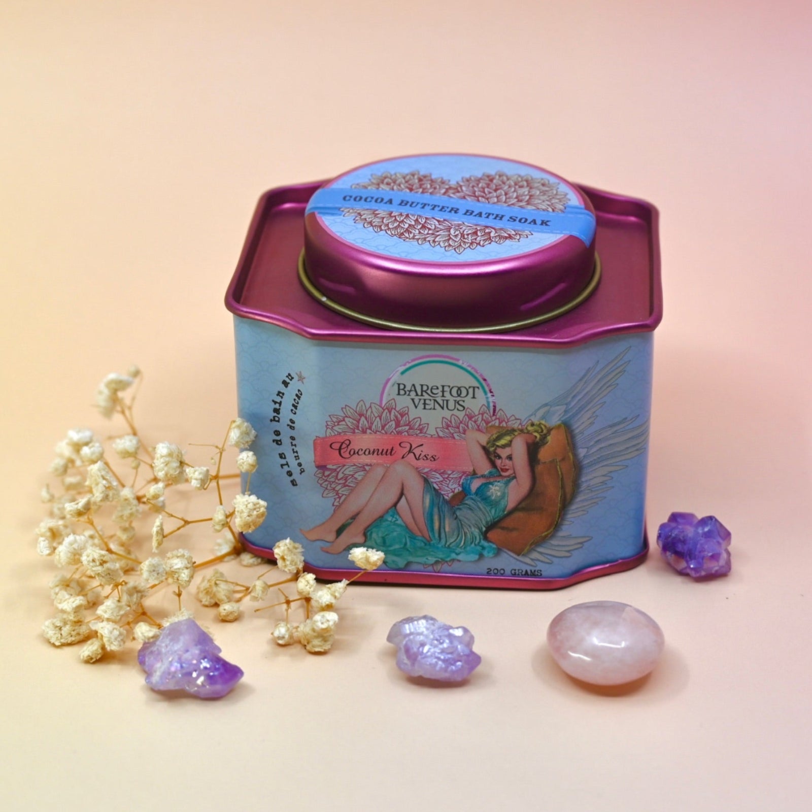 The Coconut Kiss Bath Soak surrounded by crystals and dried flowers on a light pink background.