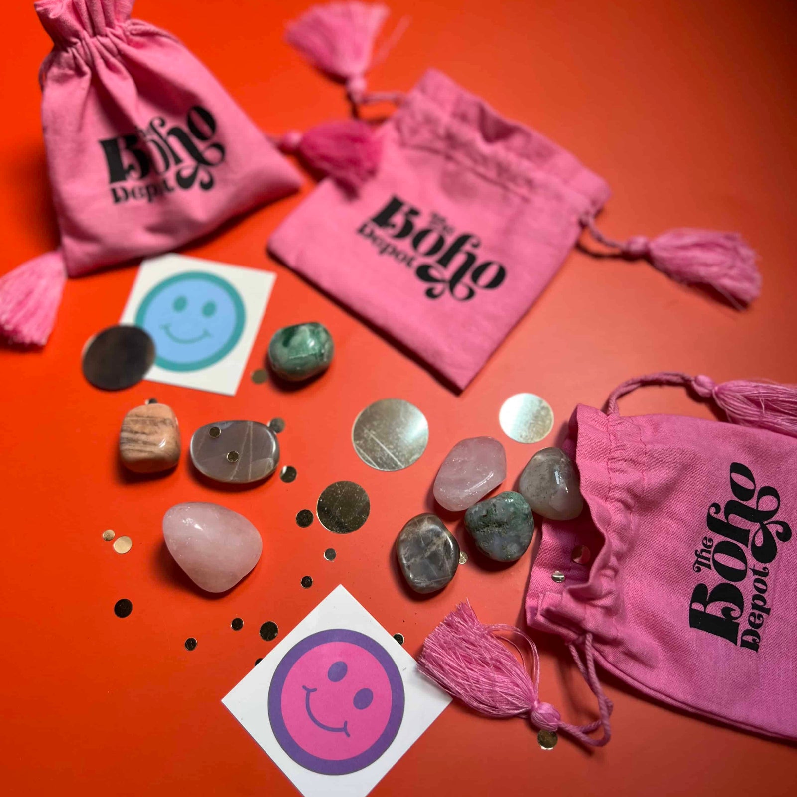 Pink drawstring bags with 'The Boho Box' branding, stones, and stickers on an orange background.