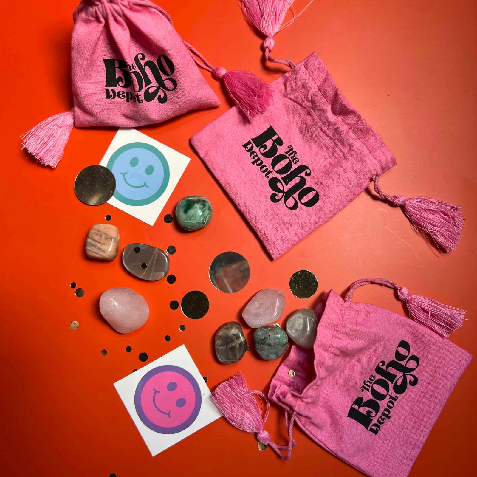 Pink drawstring bags with 'The Boho Den' branding, stones, and smiley face stickers on an orange background.