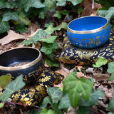 Two singing bowls, one blue and one black, on decorative stands amidst green leaves and brown leaves on the ground.