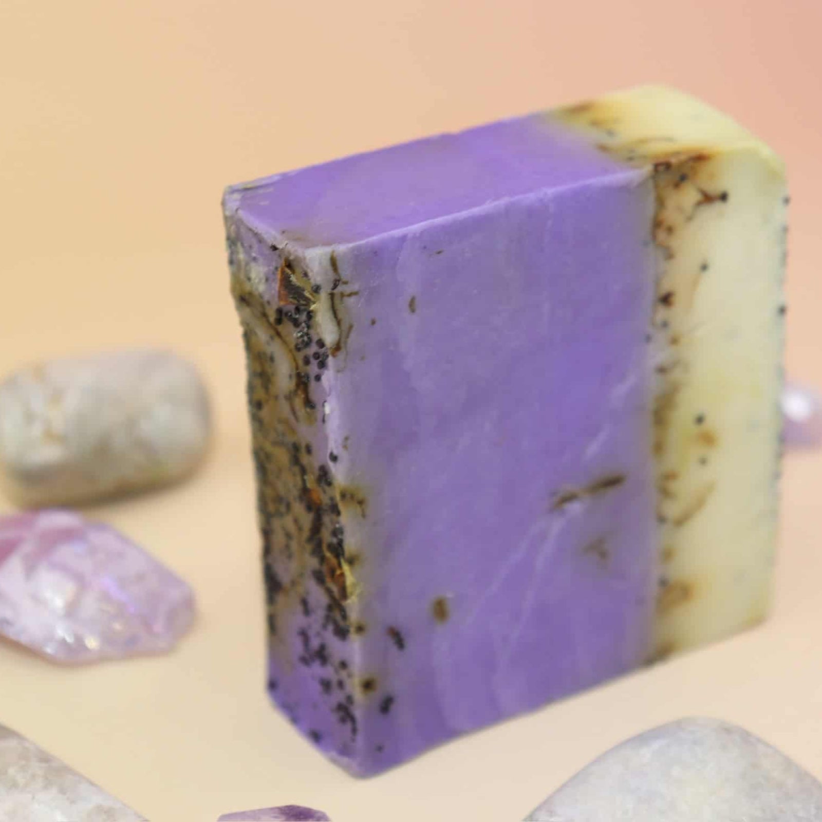 Purple soap bar with natural texture surrounded by small stones and crystals on a peach background