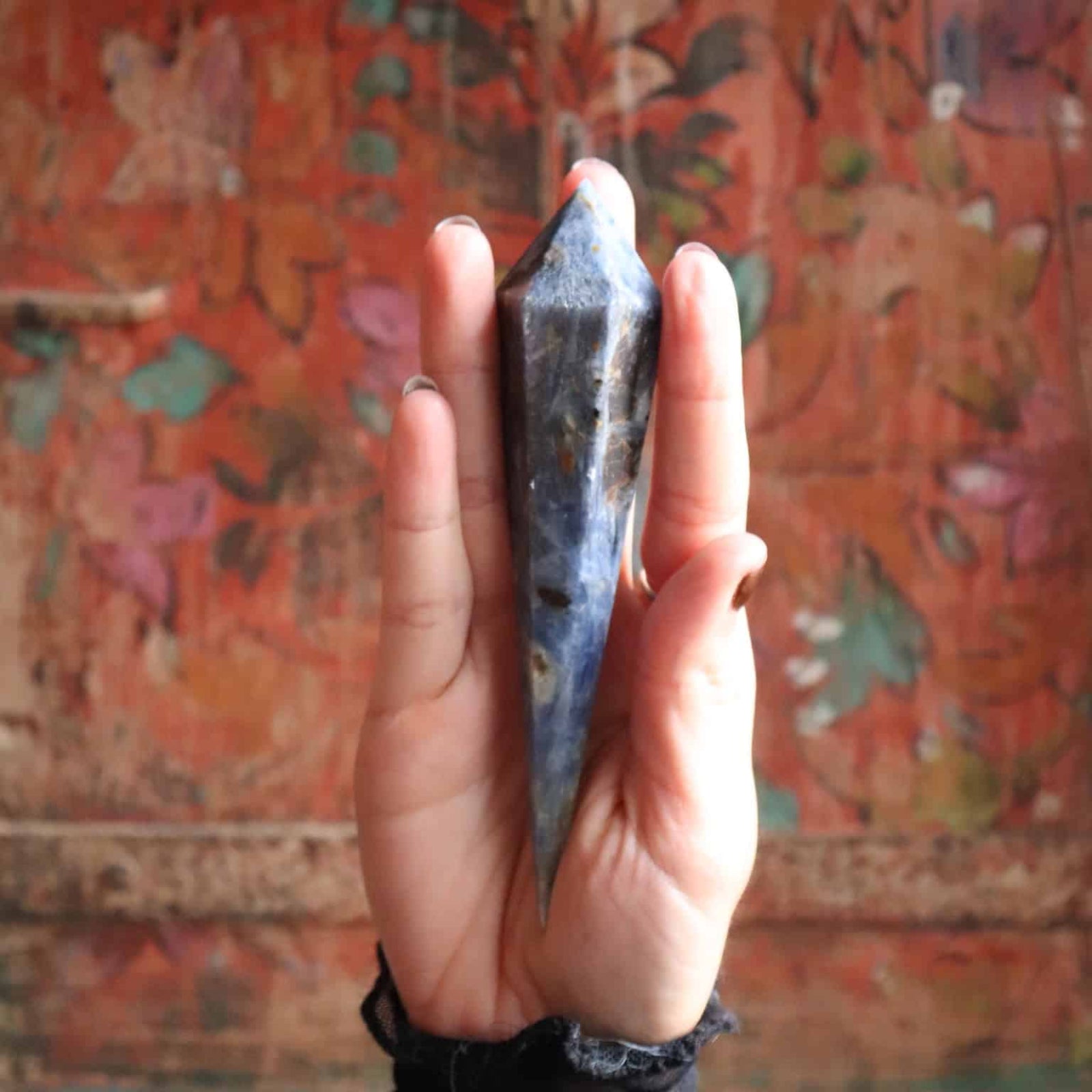 Hand holding a Blue Kyanite Scepter crystal stone in front of a colorful, patterned wall.