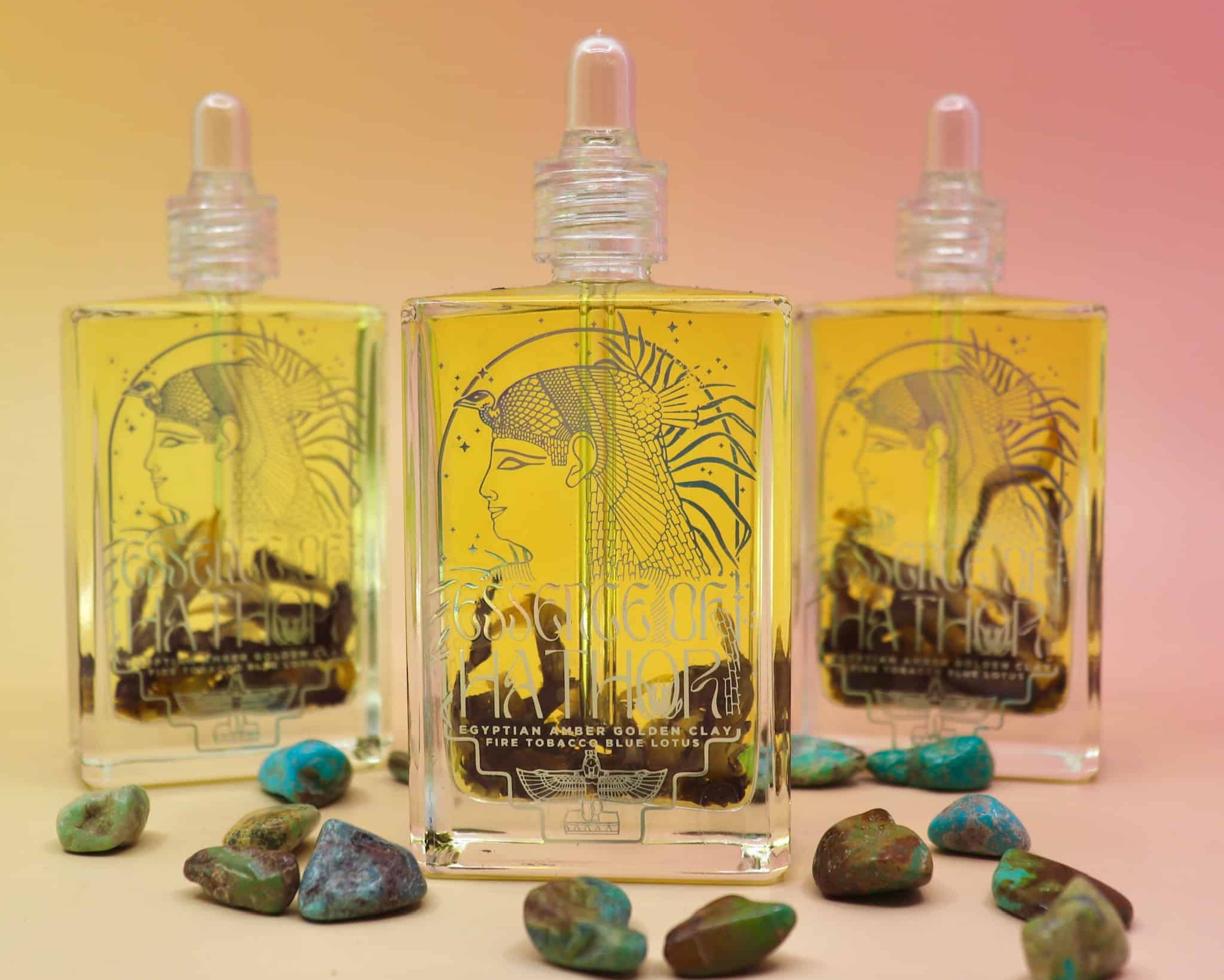 3 bottles standing up of essence of hathor blue lotus oil on a pink and orange pastel background with turquoise stones scattered in front