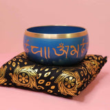 Blue singing bowl with gold text on a decorative black and gold pillow against a pink background