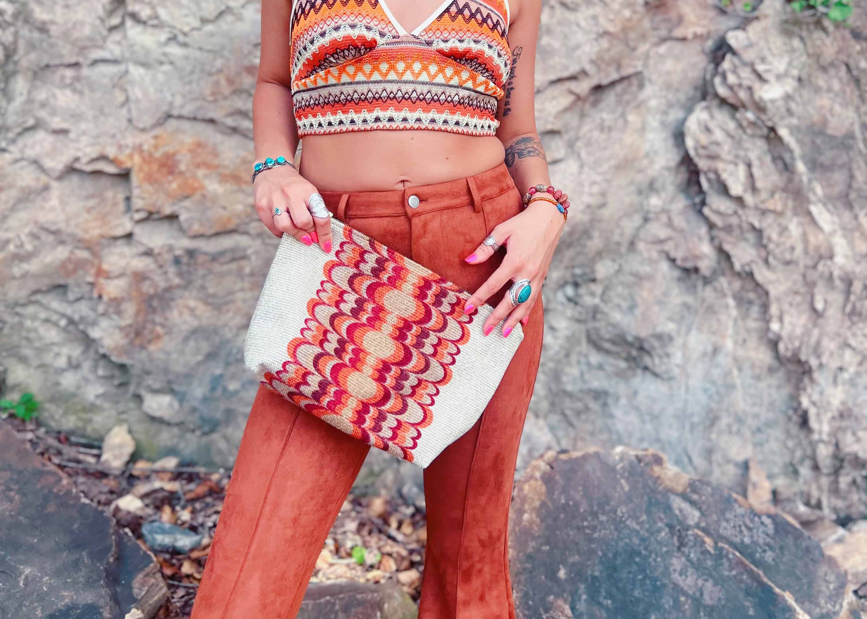 Woman in a colorful top and orange pants standing against a rocky wall.