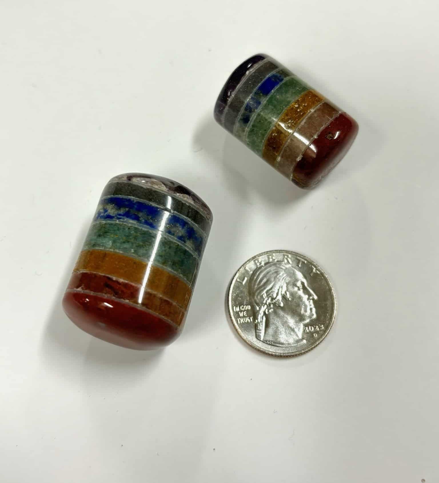 Two chakra cylinder tumbles next to a U.S. quarter to show size, featuring vibrant stacked layers of red, orange, green, blue, and purple stones
