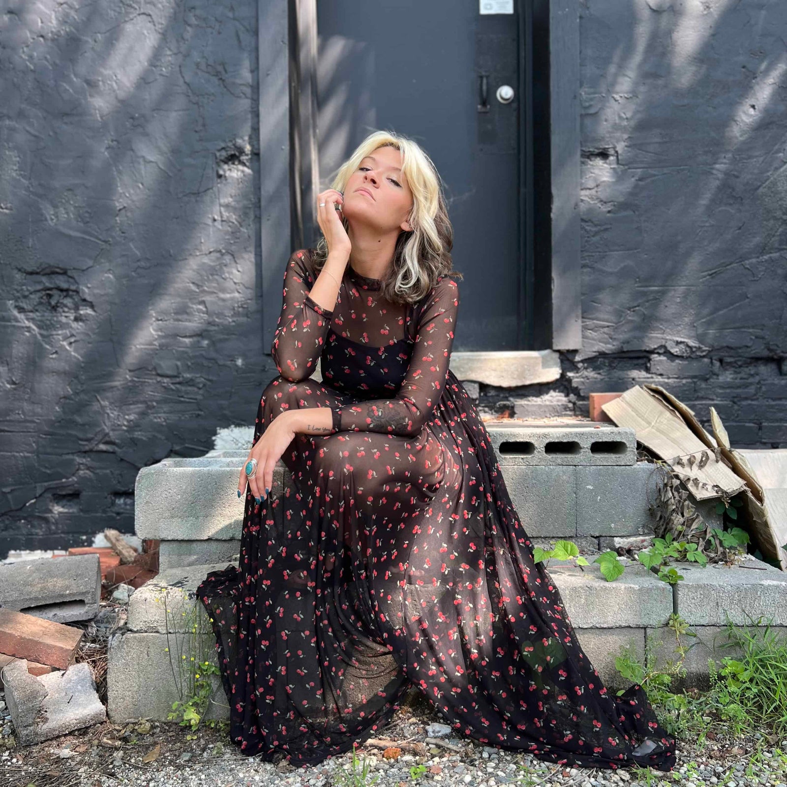 Woman seated in a cherry print mesh dress, gazing directly ahead with a soft expression.