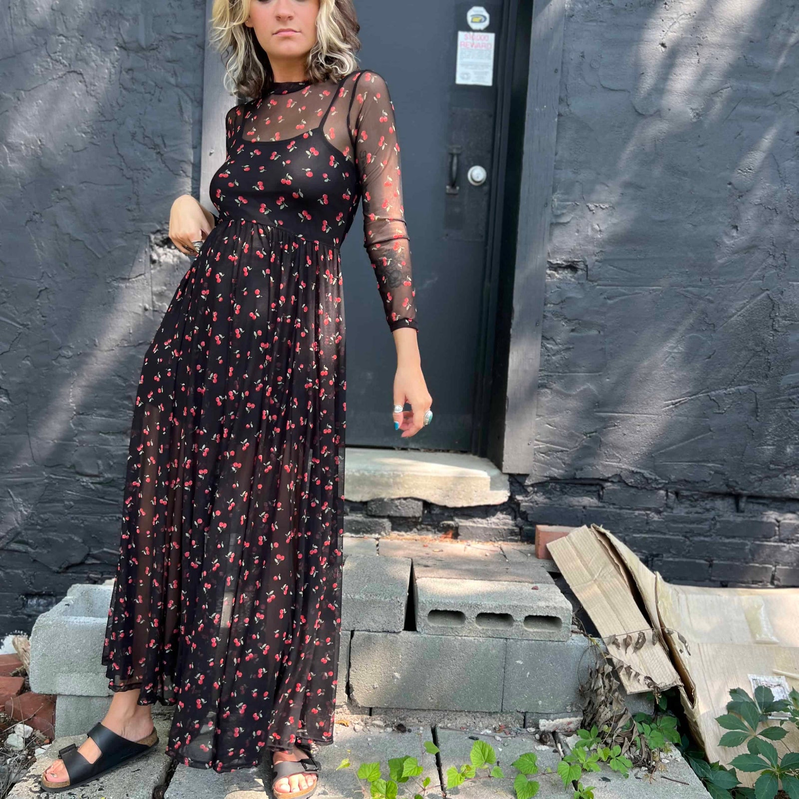 Woman in a cherry print mesh maxi dress standing on cement blocks in front of a black door
