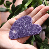 Heart-shaped Lepidolite purple stone held in a hand with green leaves in the background