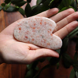 Hand holding a heart-shaped Sunstone stone with green leaves in the background