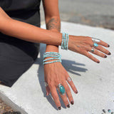 Person wearing multiple turquoise rings and larimar bracelets on a blurred outdoor background