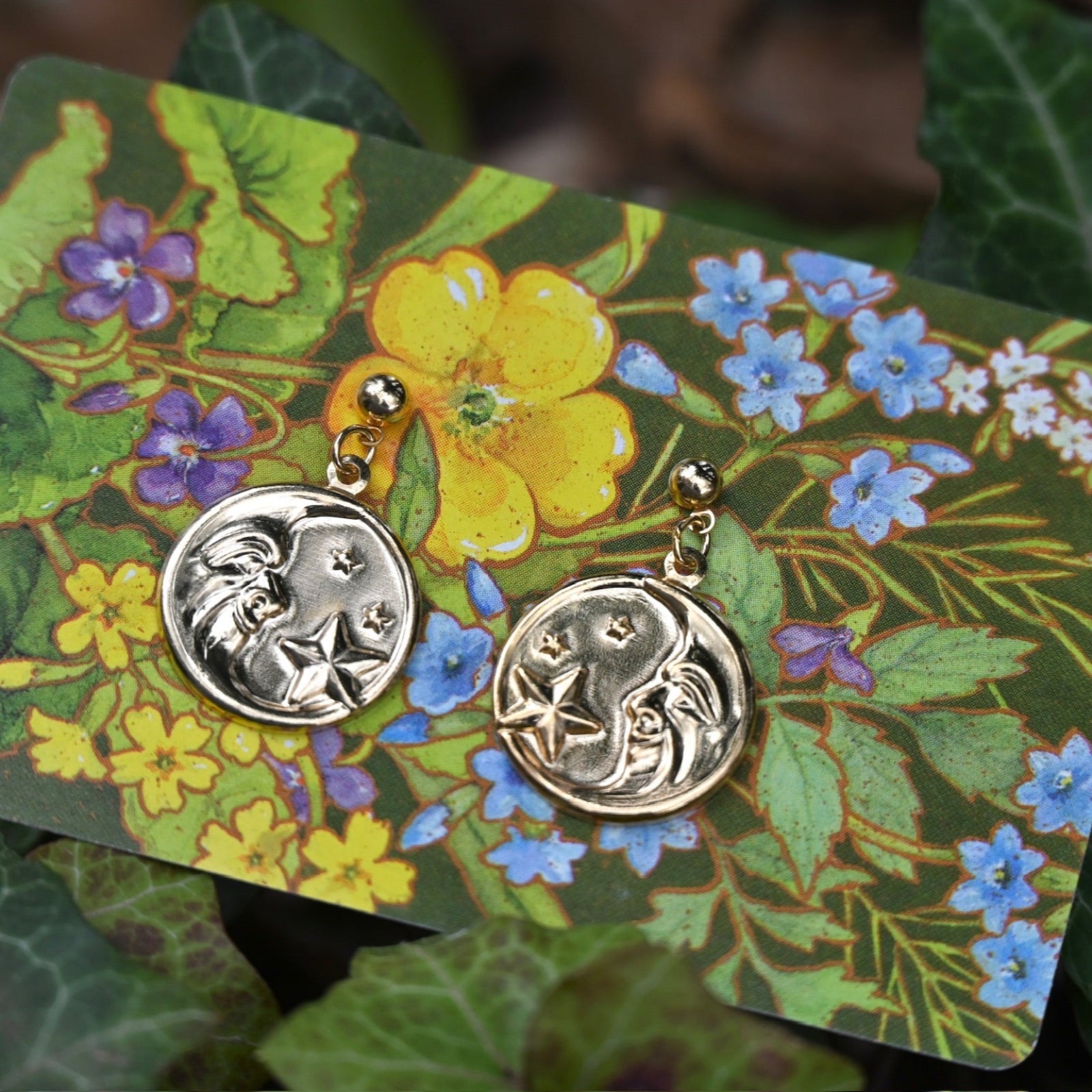 The Vintage Moon & Stars Earrings on a floral card in front of leaves.