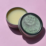 The North Country Salve on a pink background with the lid off laying slightly on top of the metal tin.
