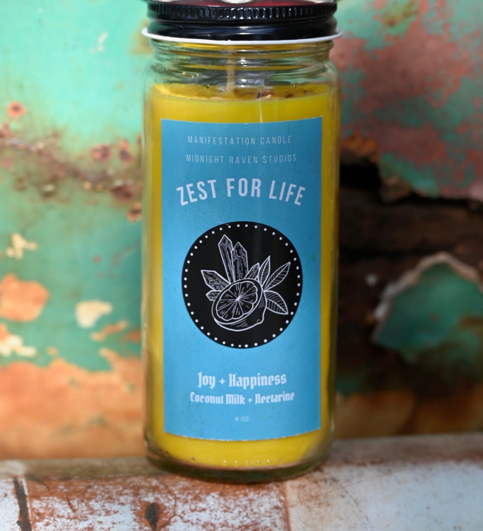 The Zest for Life Spell Candle with Herbs and Crystals in front of a teal background and on a white rusty surface.