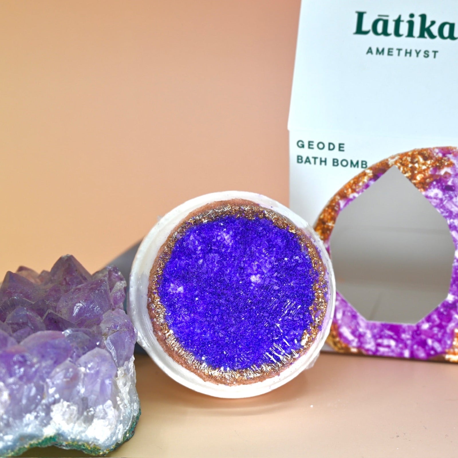The Amethyst Crystal Bath Bomb out of its box next to an amethyst piece on a pink background.