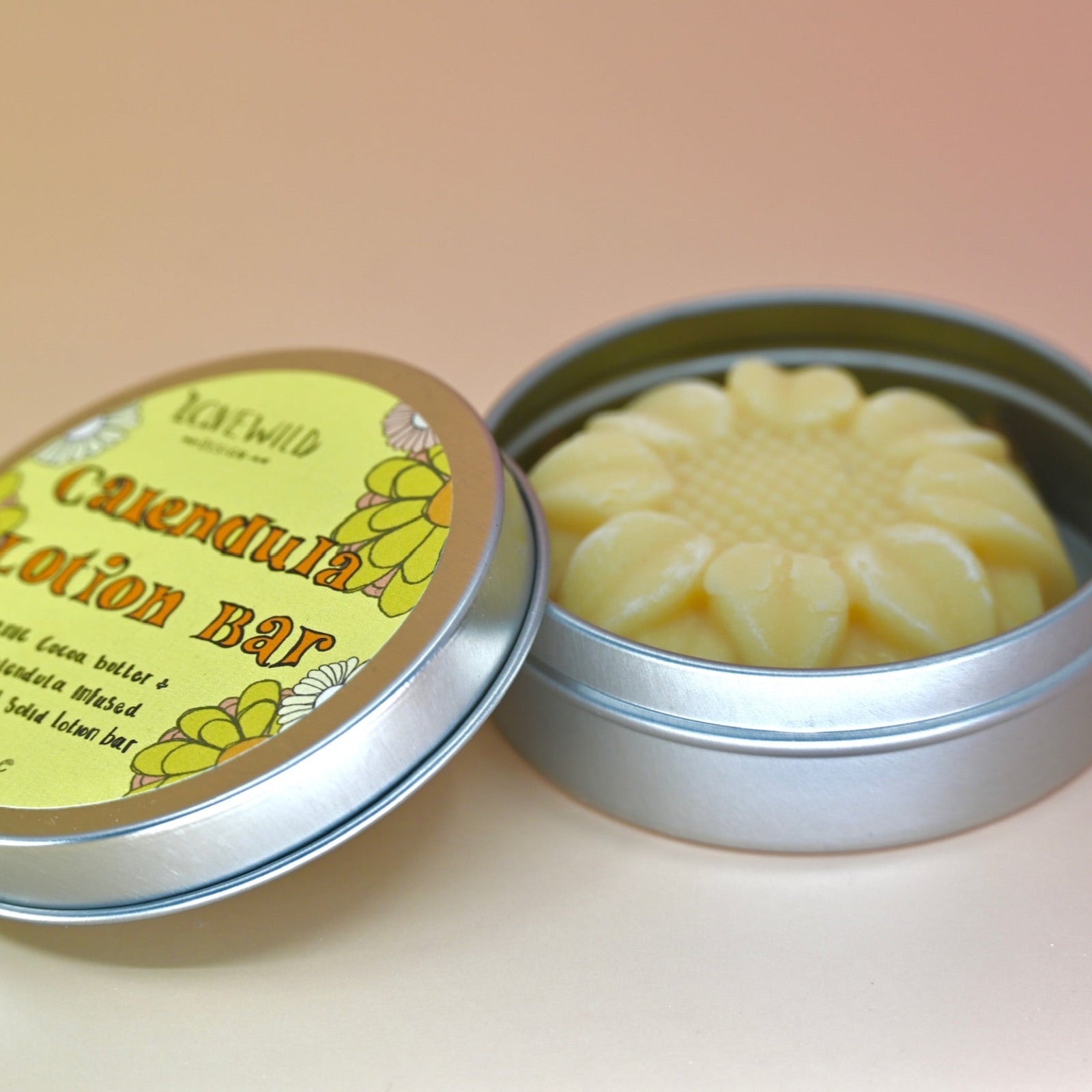 The Calendula Lotion Bar on a pink and yellow background with the tin lid off leaning against the container.