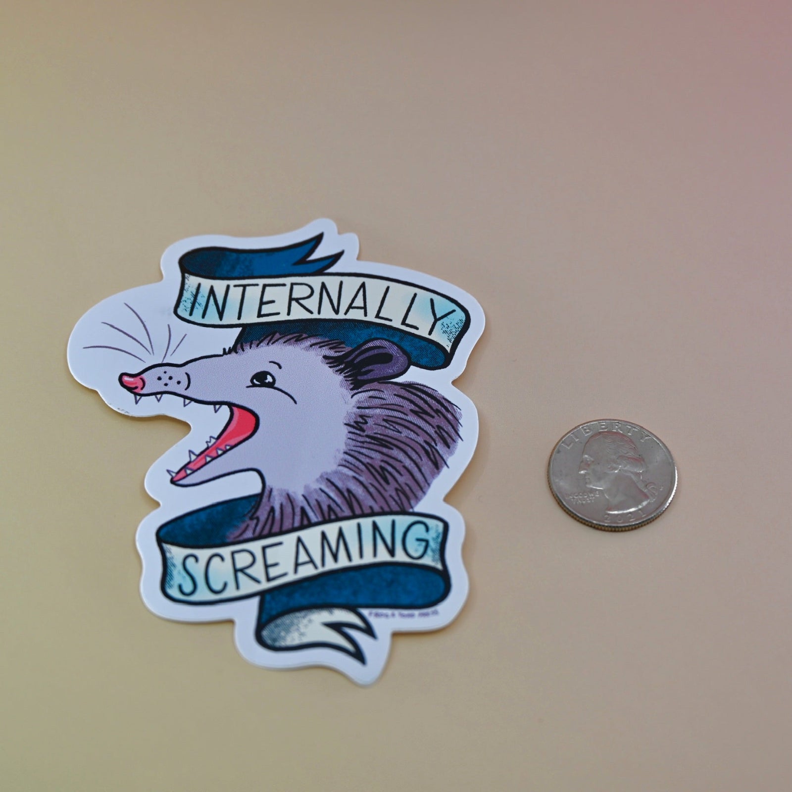 The Internally Screaming Possum Sticker next to a quarter on a tan background.