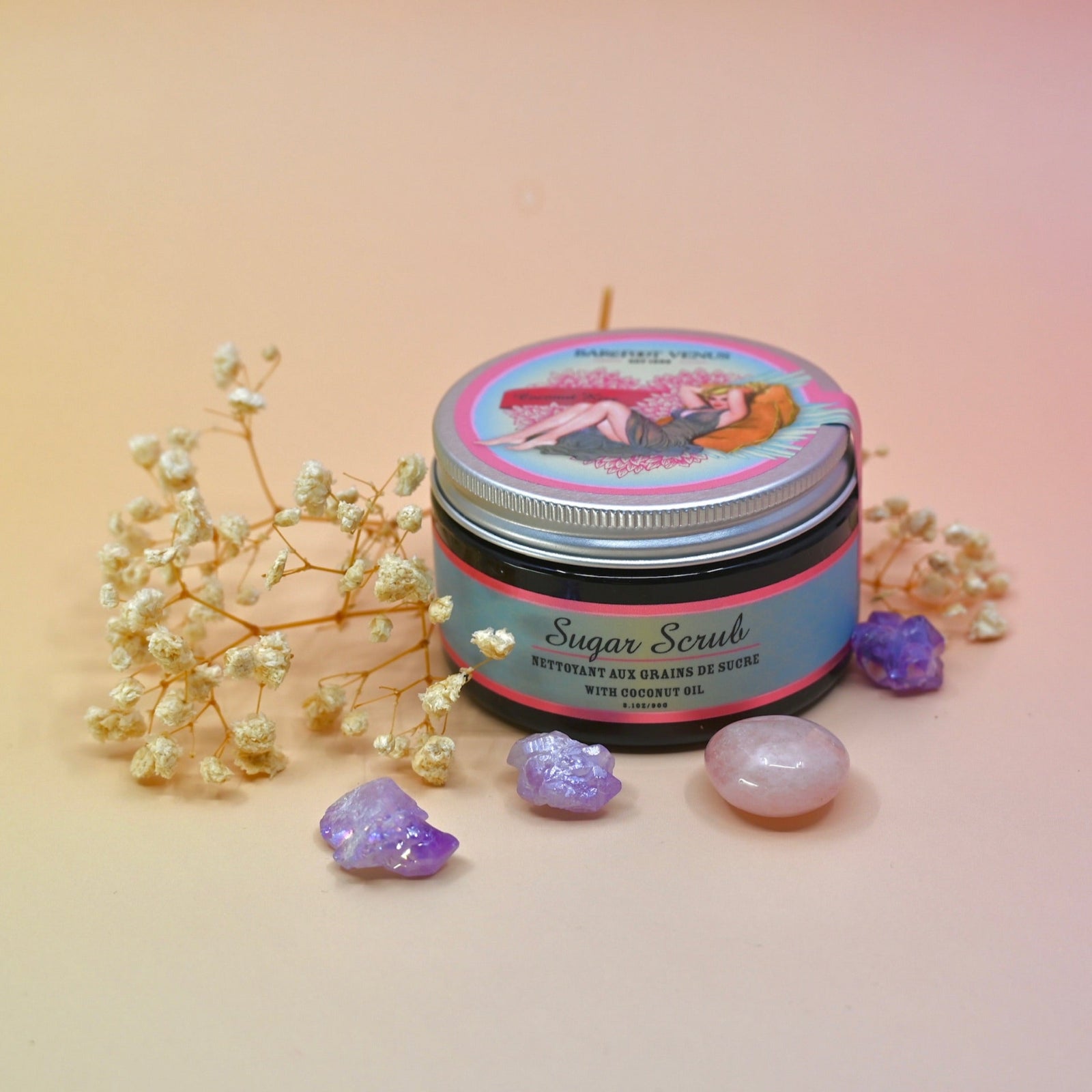 The Coconut Kiss Sugar Body Scrub surrounded by crystals and dried flowers all on a pink background.