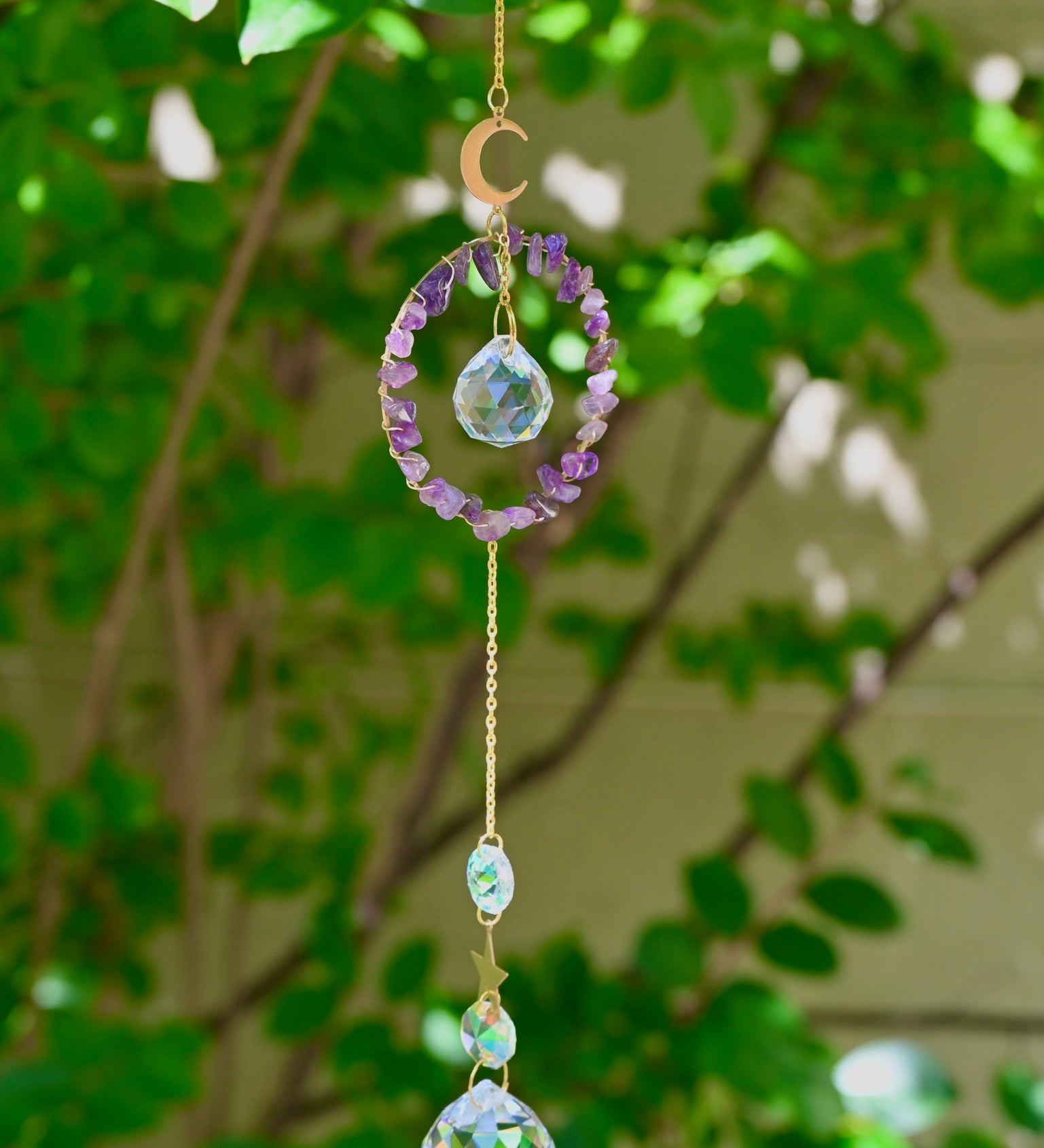 The Amethyst Gemstone Suncatcher in front of a cream-colored brick wall and greenery.