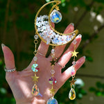 Someone holding the Opalite Crystal Moon Suncatcher with greenery in the background.