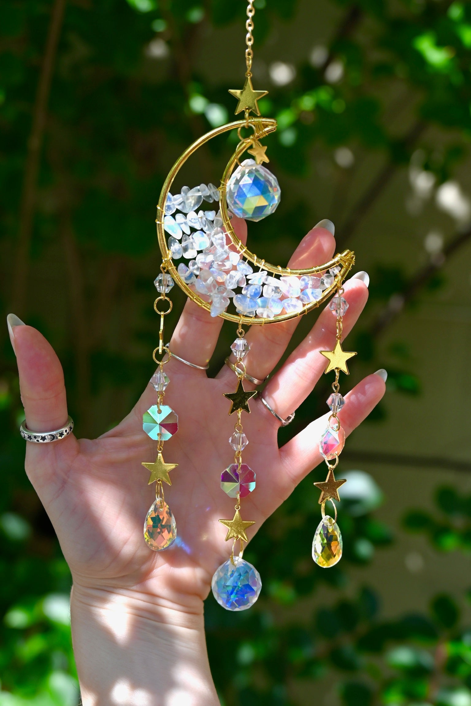 Someone holding the Opalite Crystal Moon Suncatcher with greenery in the background.