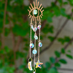 The evil eye  Fairy Lights Suncatcher in front of a cream-colored brick wall with greenery.
