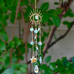 The evil eye  Fairy Lights Suncatcher in front of a cream-colored brick wall with greenery.