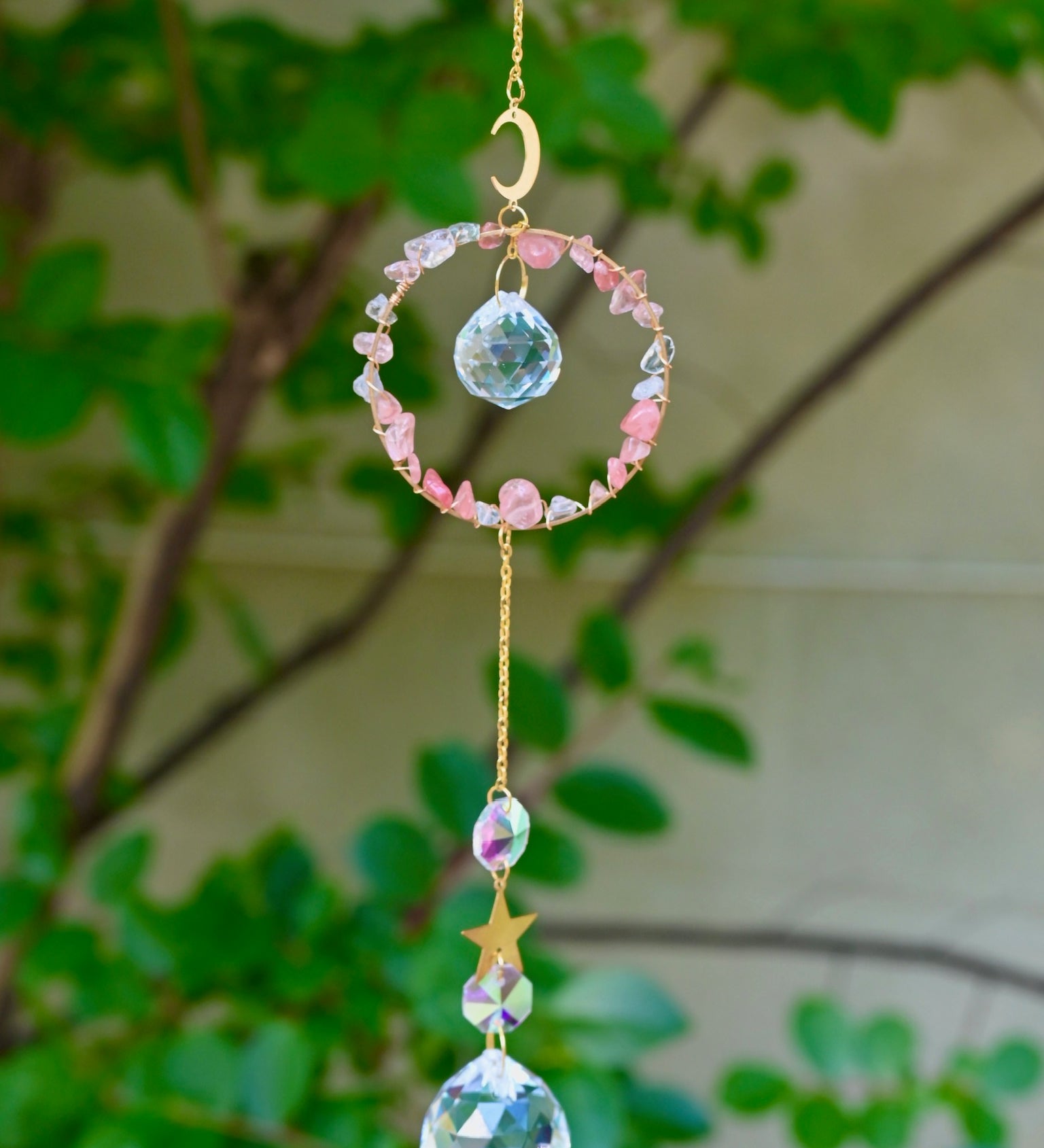 The Watermelon Quartz Gemstone Suncatcher in front of a cream-colored brick wall and greenery.