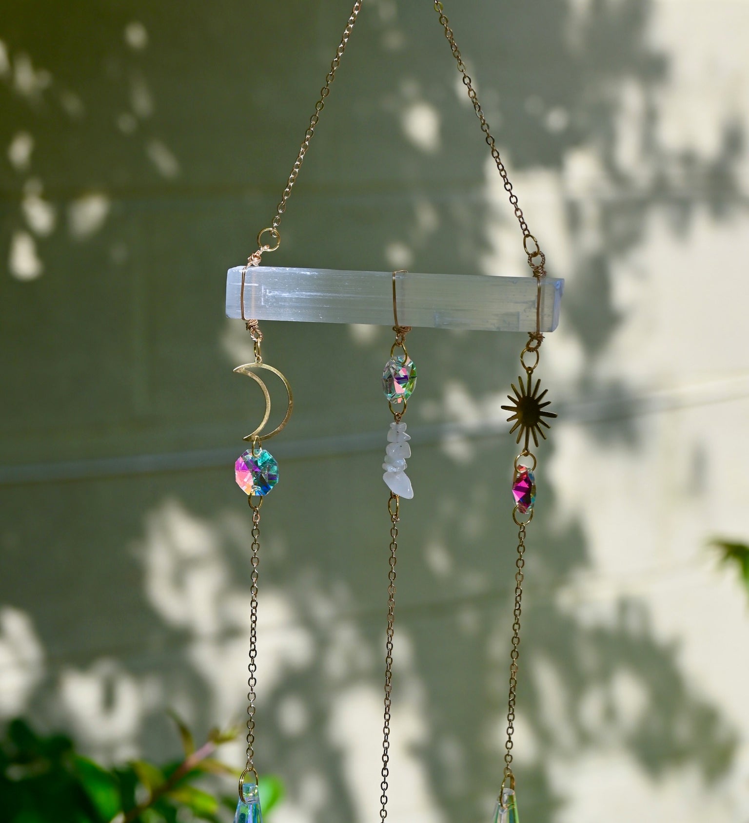 The Selenite Crystal Suncatcher in front of a white brick wall with leaves in the background.