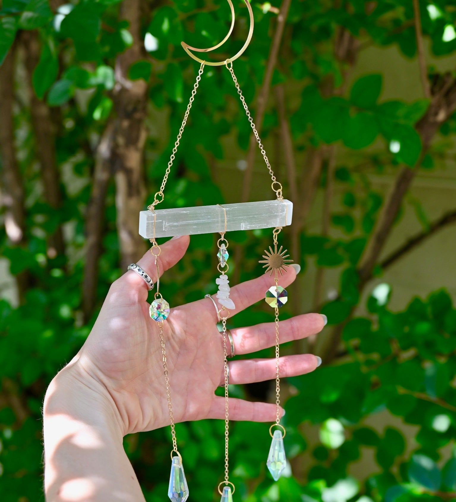 The Selenite Crystal Suncatcher in front of greenery with someone holding the gold chains.