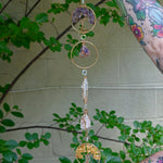 Someone holding the Chakra Gemstone Suncatcher with Amethyst, Citrine, and Quartz in front of a cream-colored brick wall with greenery.