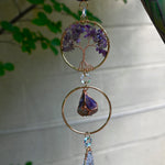 Someone holding the Chakra Gemstone Suncatcher with Amethyst, Citrine, and Quartz in front of a cream-colored brick wall with greenery.