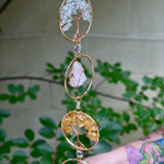 Someone holding the Chakra Gemstone Suncatcher with Amethyst, Citrine, and Quartz in front of a cream-colored brick wall with greenery.