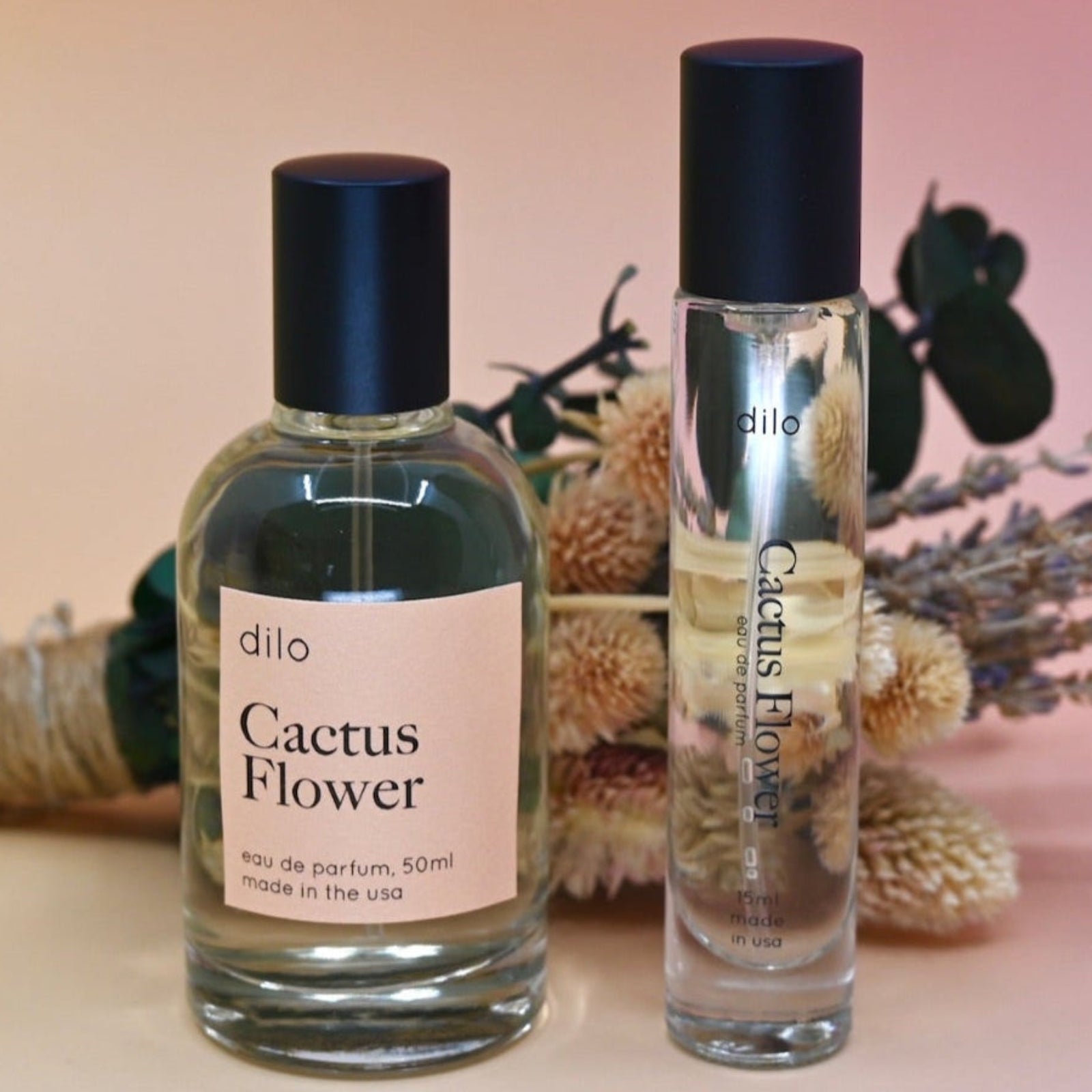 The bottle of the Cactus Flower 15ml Unisex Eau de Parfum Travel Sprayer and regular sized bottle on a pink background in front of a dried flower bundle.