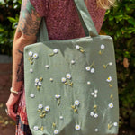 Blonde woman wearing a floral dress and carrying a sage green linen tote bag with hand-embroidered white daisies