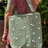 Blonde woman wearing a floral dress and carrying a sage green linen tote bag with hand-embroidered white daisies