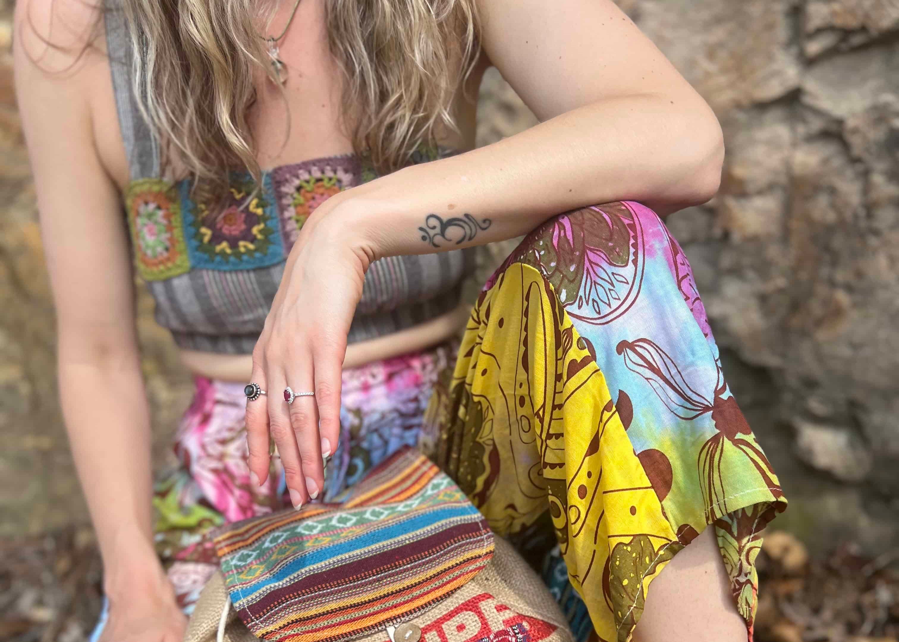 Person wearing a colorful outfit with a patterned bag sitting against a stone wall.