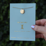 Someone holding the Gemini 14k Gold Dipped Nickel-Free Zodiac Necklace on its card in front of greenery.