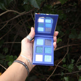 Someone holding the Healing Waters 6 Pan Vegan Eyeshadow Palette open in front of a bush, showing the inside.