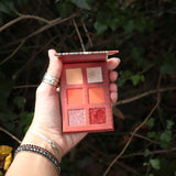 Someone holding the Sun Fire 6 Pan Vegan Eyeshadow Palette open in front of a green bush.