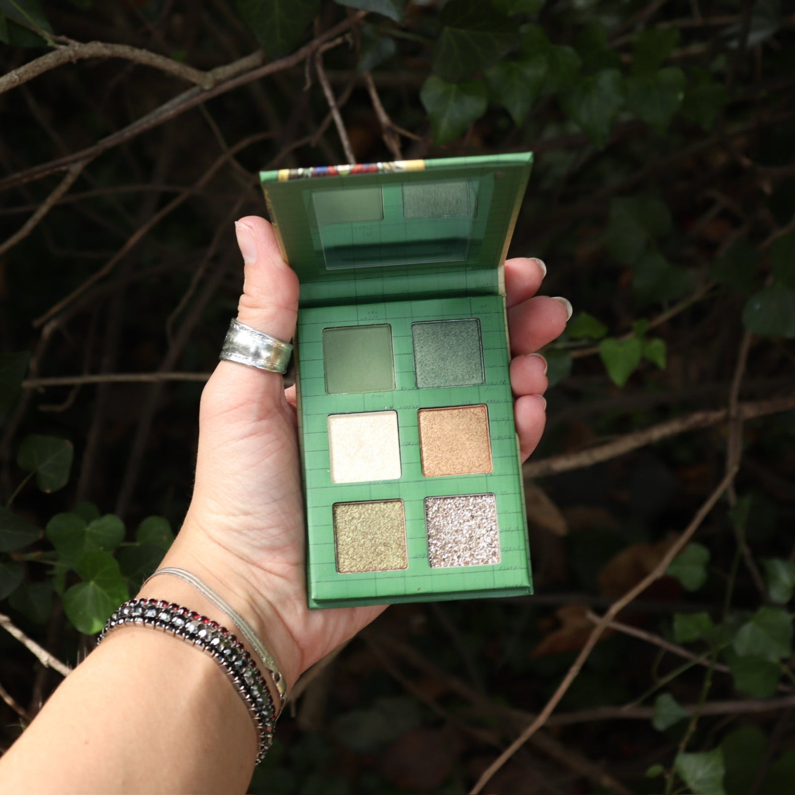 Someone holding the Plant Medicine 6 Pan Vegan Eyeshadow Palette open, in front of a green bush.