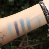 Someone wearing test swabs of the Healing Waters 6 Pan Vegan Eyeshadow Palette on their arm in front of a green bush.