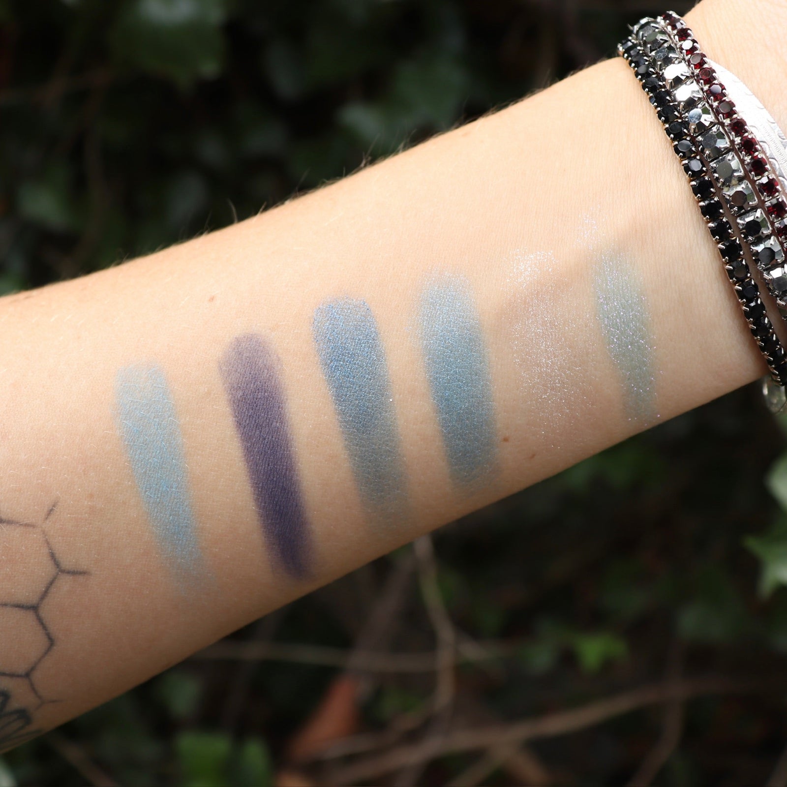 Someone wearing test swabs of the Healing Waters 6 Pan Vegan Eyeshadow Palette on their arm in front of a green bush.