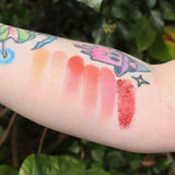 Someone with test swabs of the colors from the Sun Fire 6 Pan Vegan Eyeshadow Palette on their arm in front of a green bush.