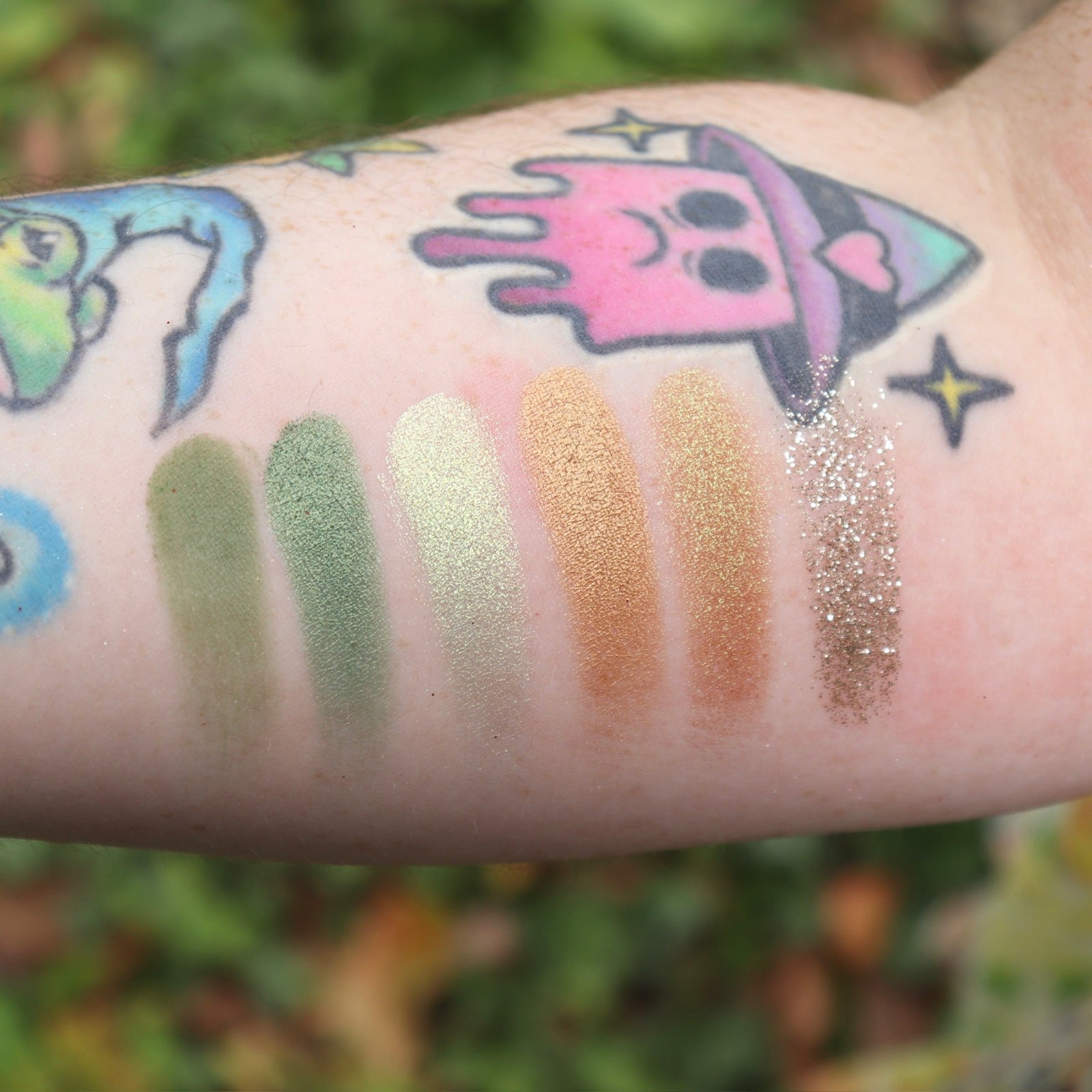 Someone wearing test swabs on their arm of the Plant Medicine 6 Pan Vegan Eyeshadow Palette in front of a green bush.