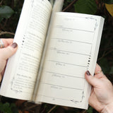Someone holding The Practical Witch's 2026 Almanac open in front of some greenery.