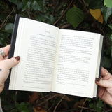 Someone holding the Dictionary of Obscure Sorrows Hardcover Book open in front of a green bush, showing off the pages.