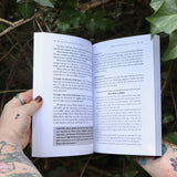 Someone holding the Sacred Herbs of Yule and Christmas open in front of a green bush, showing off the pages.