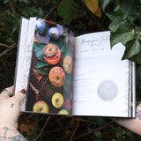 Someone holding the Natural Witch's Cookbook Hardcover Book open in front of a green bush, showing off the pages.