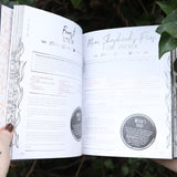 Someone holding the Natural Witch's Cookbook Hardcover Book open in front of a green bush, showing off the pages.